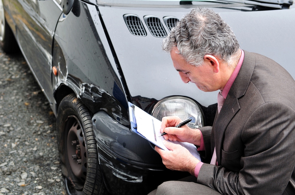 Insurance representative writing on clipboard while looking at damaged car after collision