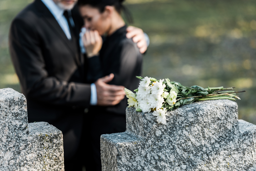 Flowers on tombstone, man comforting woman in background